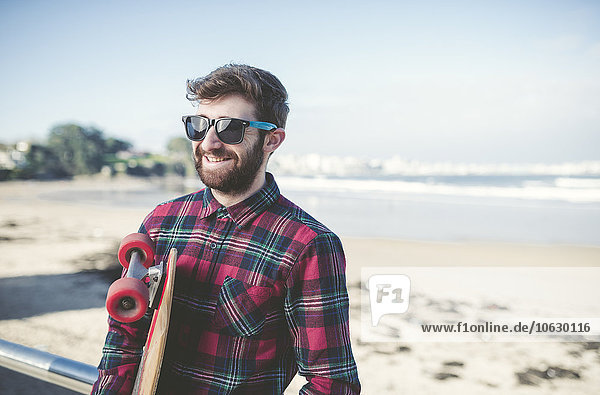 Spanien  La Coruna  Porträt eines lächelnden Hipsters mit Sonnenbrille und Skateboard am Strand