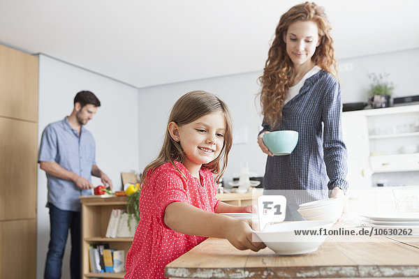 Smiling little girl laying the table in the kitchen with her parents in the background