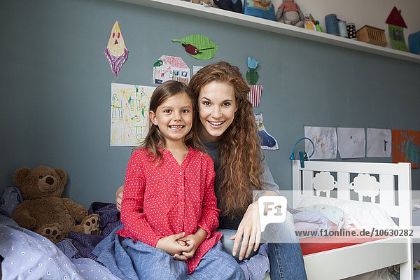 Porträt der Mutter und der kleinen Tochter die nebeneinander auf dem Bett im Kinderzimmer sitzen.