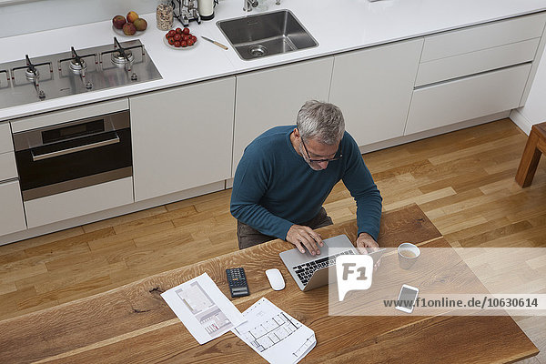 Reifer Mann in der Küche mit Bauplan mit Laptop