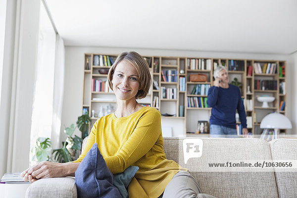 Lächelnde Frau sitzt auf der Couch, während ihr Mann im Hintergrund telefoniert.