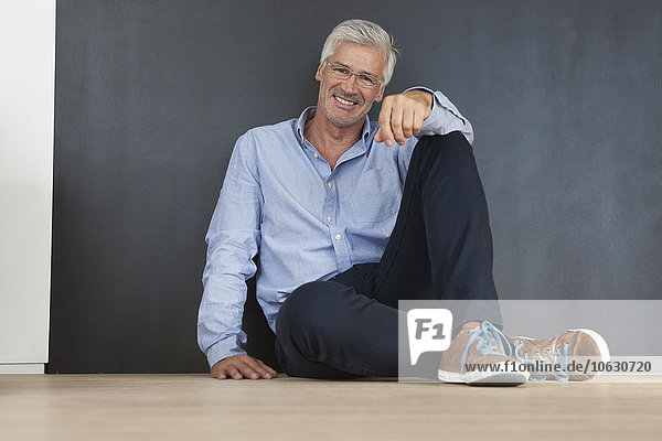 Porträt eines lächelnden reifen Mannes, der auf dem Boden vor einer grauen Wand sitzt