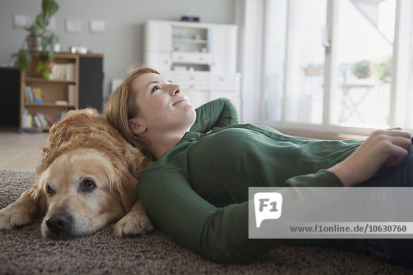 Lächelnde junge Frau liegt mit ihrem Hund auf dem Teppich zu Hause.