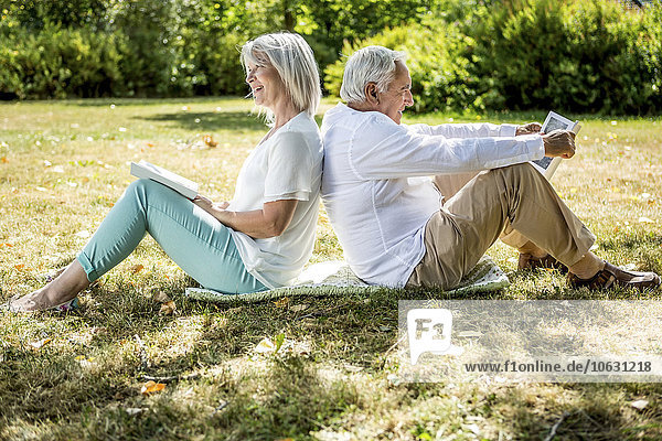 Elderly couple sitting back to back in grass reading books