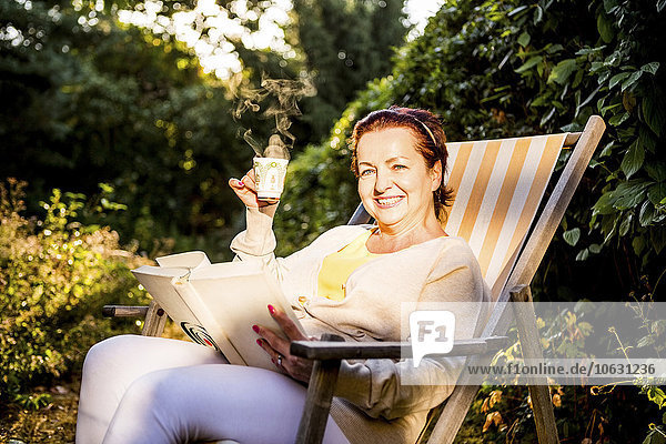 Smiling mature woman reading book and drinking coffee in deck chair in garden