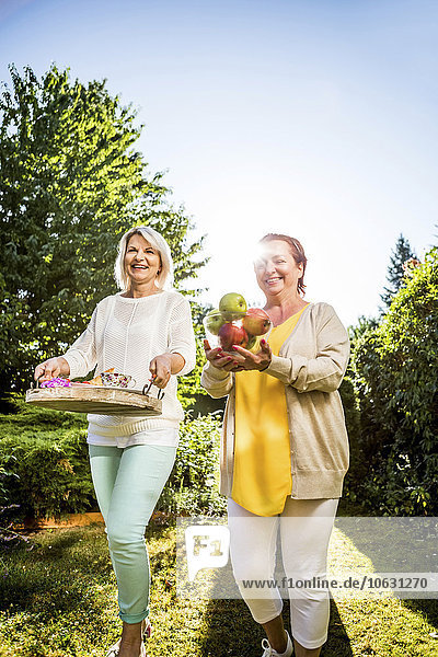 Two happy mature women carrying apples and tray in garden