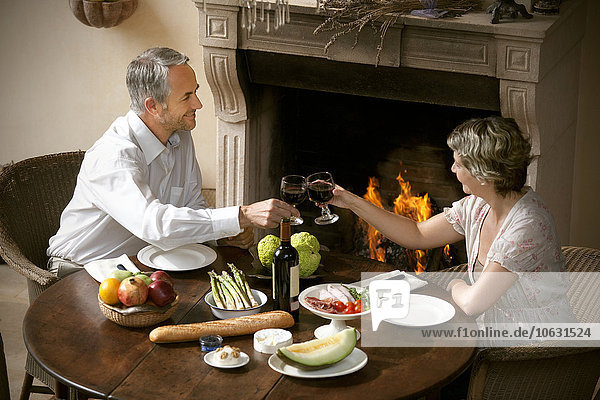 Ein reifes Paar sitzt am gedeckten Tisch und toastet mit Rotwein vor offenem Feuer.