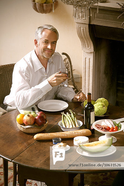 Portrait of smiling mature man sitting at laid table in his living room holding glass of red wine