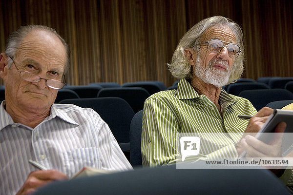 Two senior men sitting in auditorium taking notes