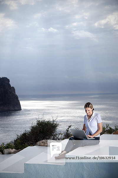 Spanien  Mallorca  Frau mit Laptop auf der Treppe sitzend mit dem Meer im Hintergrund
