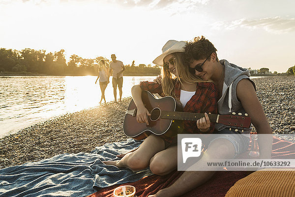 Junges Paar mit Gitarre entspannt am Flussufer bei Sonnenuntergang