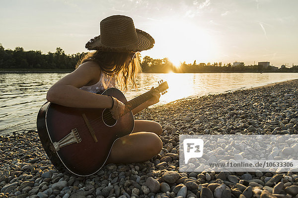 Young woman playing guitar at the riverside at sunset