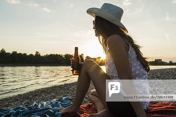 Young woman drinking beer at the riverside at sunset