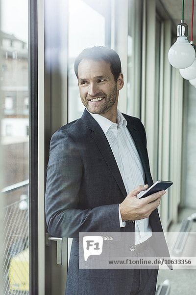 Porträt eines lächelnden Geschäftsmannes mit Smartphone durchs Fenster schauend