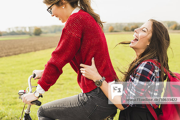 Zwei glückliche junge Frauen teilen sich ein Fahrrad in ländlicher Landschaft.