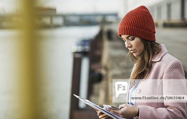 Junge Frau am Flussufer schaut auf Handy und Dokumente