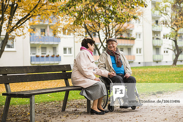 Seniorin auf der Bank neben dem Ehemann im Rollstuhl sitzend