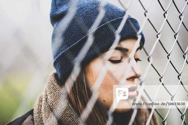 Portrait of young woman with closed eyes wearing wool cap behind wire mesh