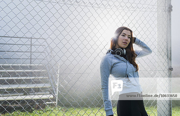 Porträt einer jungen Frau in Sportbekleidung  die sich an einen Gitterzaun lehnt.