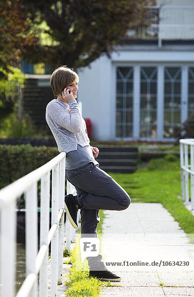 Österreich  Mondsee  lächelnde junge Frau lehnt sich an das Geländer und telefoniert mit dem Smartphone.