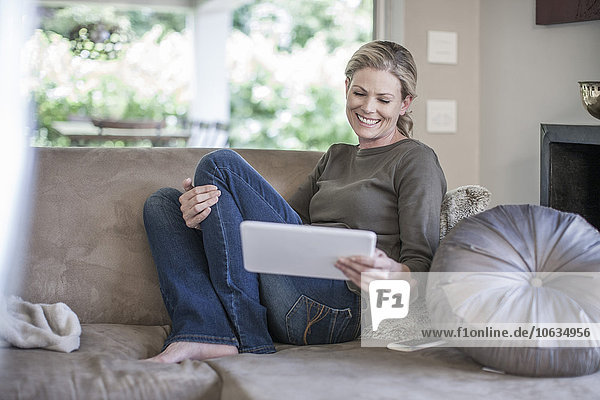 Lächelnde Frau sitzt auf der Couch und schaut auf ein digitales Tablett.