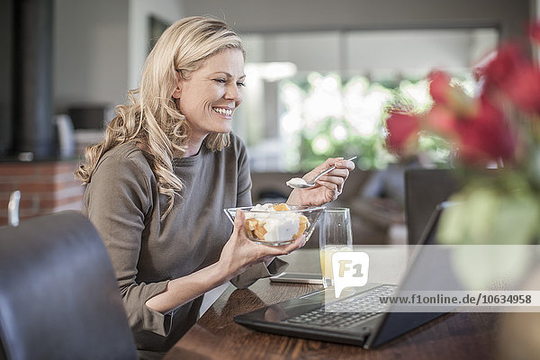 Frau isst Obstsalat und arbeitet zu Hause am Laptop