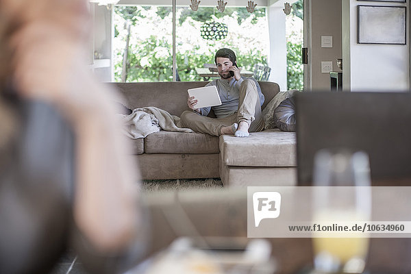 Mann auf der Couch beim Betrachten des digitalen Tabletts und beim Telefonieren mit der Frau im Vordergrund