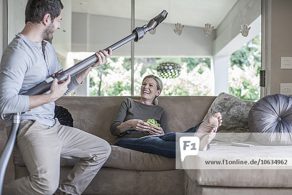 Frau auf der Couch und Mann  der vorgibt  mit dem Staubsauger Gitarre zu spielen.