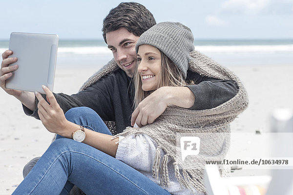 Ein Paar sitzt am Strand und nimmt einen Selfie mit digitalem Tablett.