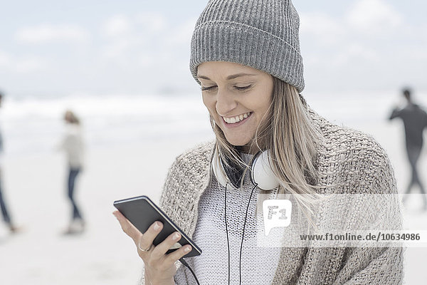 Portrait of smiling woman wearing beanie looking at smartphone on the beach