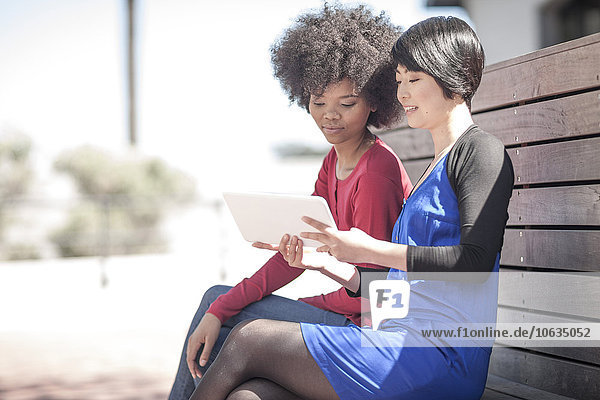 Two young women sitting on a bench looking at digital tablet