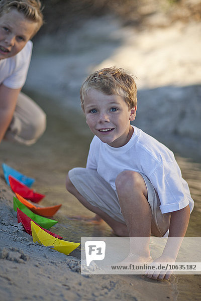 Zwei Jungen spielen mit Papierbooten am Strand.