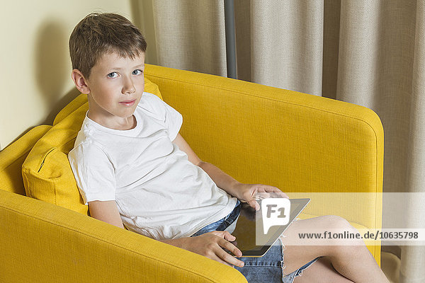 Porträt eines Jungen mit digitalem Tablett  der zu Hause auf einem Sessel sitzt.