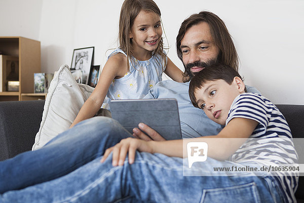 Vater mit Kindern  die zu Hause ein digitales Tablett auf dem Sofa benutzen
