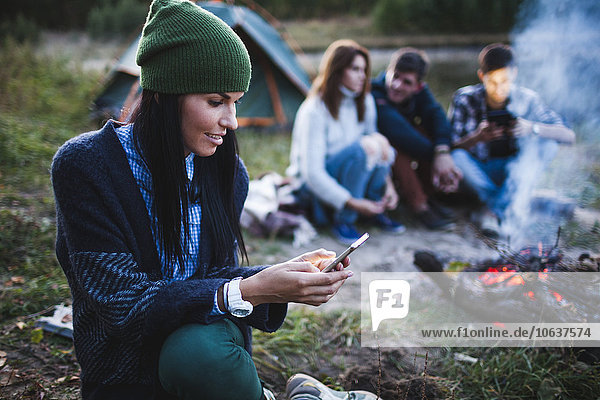 Junge Frau mit Handy  während Freunde am Lagerfeuer auf dem Campingplatz sitzen