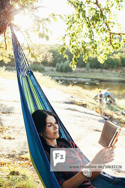 Junge Frau beim Lesen eines Buches auf der Hängematte im Wald