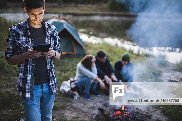 Young man using digital tablet while friends sitting by bonfire in forest