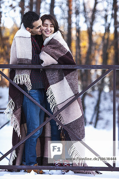 Full length of young man kissing woman wrapped in blanket on bridge during winter