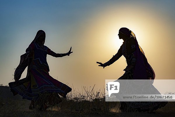Two young women in dresses dancing in front of the setting sun  Pushkar Camel Fair  Pushkar  Rajasthan  India  Asia