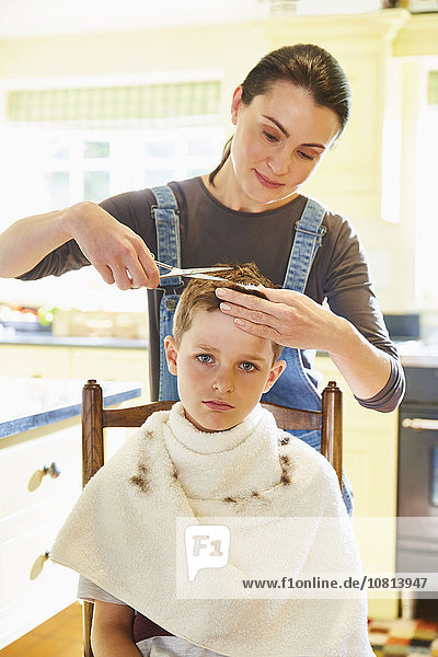 Porträt eines unglücklichen Jungen, der sich von seiner Mutter in der Küche die Haare schneiden lässt.