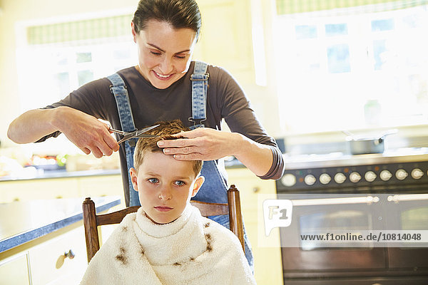 Unhappy boy getting haircut from mother in kitchen