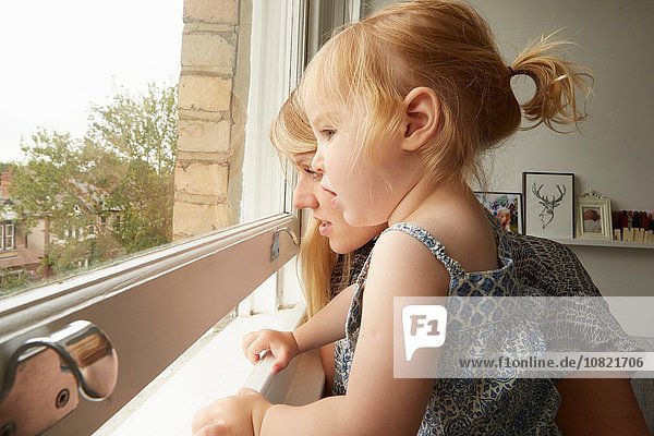 Mid adult woman and toddler daughter gazing out from living room window