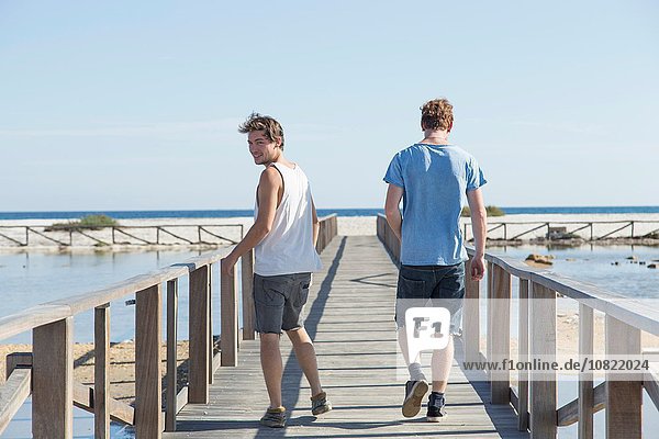 Durchgehende Rückansicht von jungen Männern  die auf einem hölzernen Pier laufen und über die Schulter schauen  Sardinien  Italien
