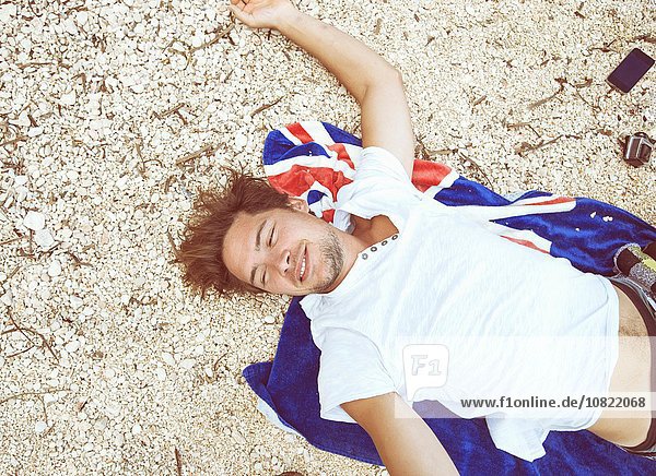 Overhead-Ansicht eines jungen Mannes  der am Strand auf einem Strandtuch liegt und lächelnd wegsieht  Costa Smeralda  Sardinien  Italien