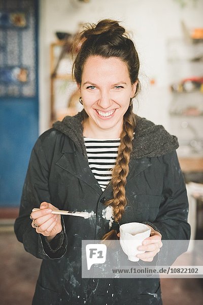 Portrait of young woman holding clay pot and paintbrush looking at camera smiling