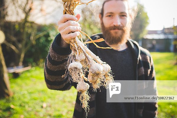 Ein bärtiger Mann im Garten mit frisch gepflückten Knoblauchzwiebeln und lächelndem Blick auf die Kamera.