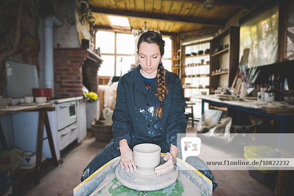 Front view of young woman in workshop sitting at pottery wheel making clay pot  looking down