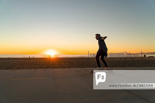 Young man using skateboard on pathway  beside beach  sunset