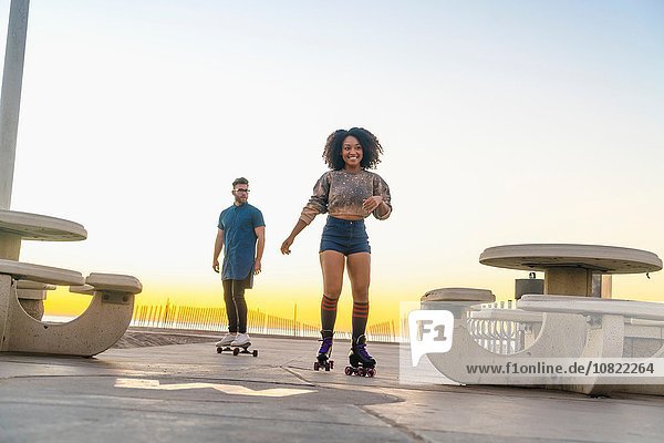 Young man on skateboard  following mid adult woman on rollerskates