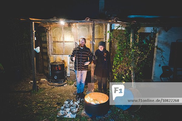 Couple in front of shed holding tongs  with pottery by fire in barrel